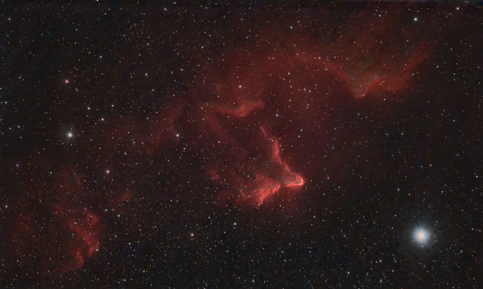 Red glowing emission nebula with a bright rim, shaped like a ghost, in a star field with a bright blue-white star at lower right